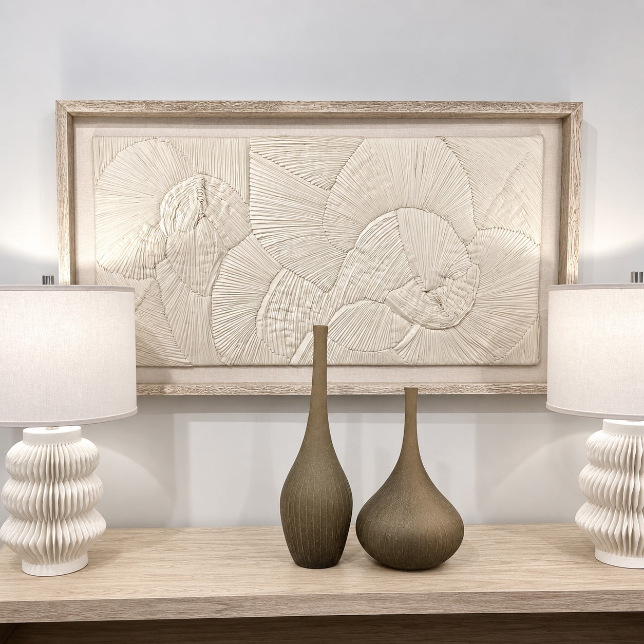 A large rectangular framed wall art piece with a textured, embossed floral or sunburst design in warm off-white tones, displayed above a console table with two white ribbed table lamps and two brown vases.
