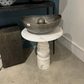 Decorative metal bowl on a marble pedestal in a room setting with other decorative items.