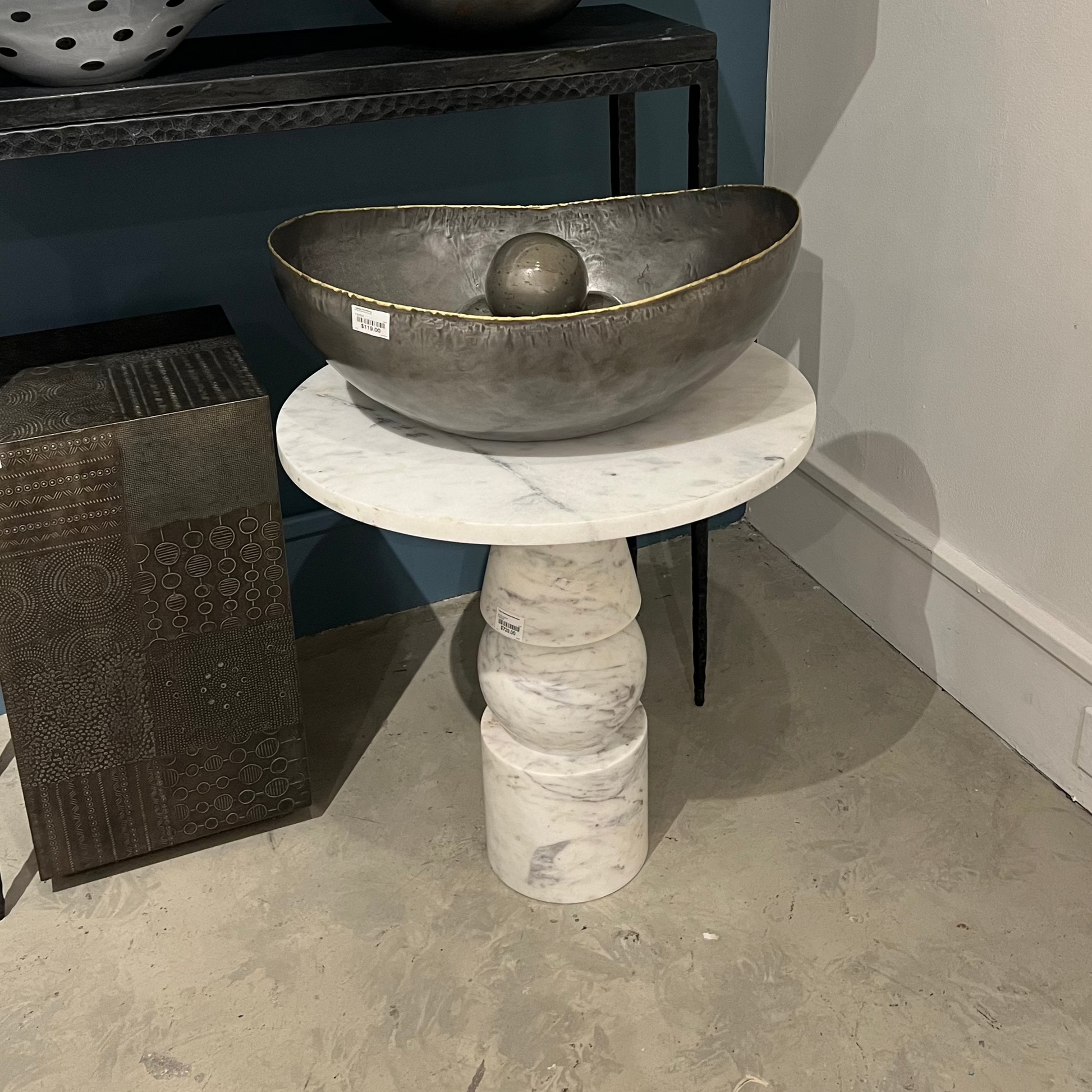Decorative metal bowl on a marble pedestal in a room setting with other decorative items.
