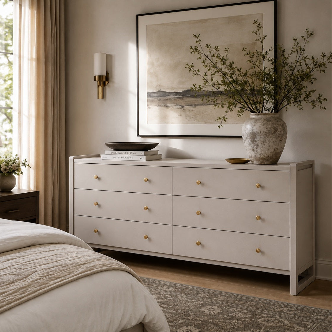 A light beige six-drawer dresser in a styled bedroom setting, decorated with a large ceramic vase of branches, books, and a bowl, beneath a framed landscape print.