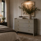 A styled bedroom interior featuring a light gray three-drawer dresser with small gold knobs, styled with books, a vase of greenery, and a framed print on a beige wall. A bed with neutral linens and a dark throw blanket is visible on the left, along with a bedside lamp and window.