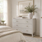 A light gray three-drawer dresser with small gold knobs in a bright, neutral bedroom. The dresser is styled with a textured ceramic vase containing greenery, a stack of books, and a framed print on the wall above it.