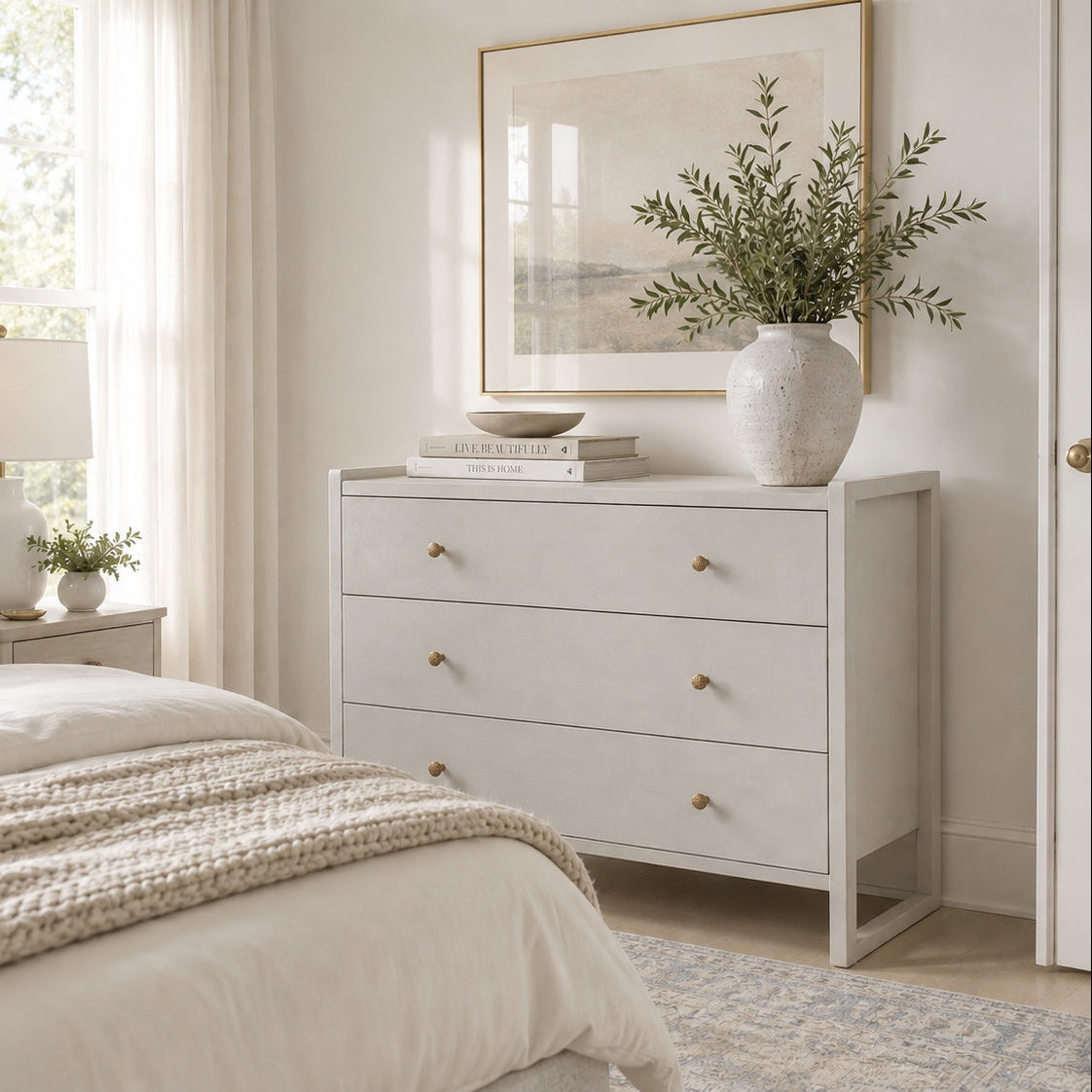 A light gray three-drawer dresser with small gold knobs in a bright, neutral bedroom. The dresser is styled with a textured ceramic vase containing greenery, a stack of books, and a framed print on the wall above it.