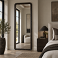 A tall floor mirror with an ornate dark frame stands in a styled bedroom next to a bed and nightstand, reflecting a softly lit interior.