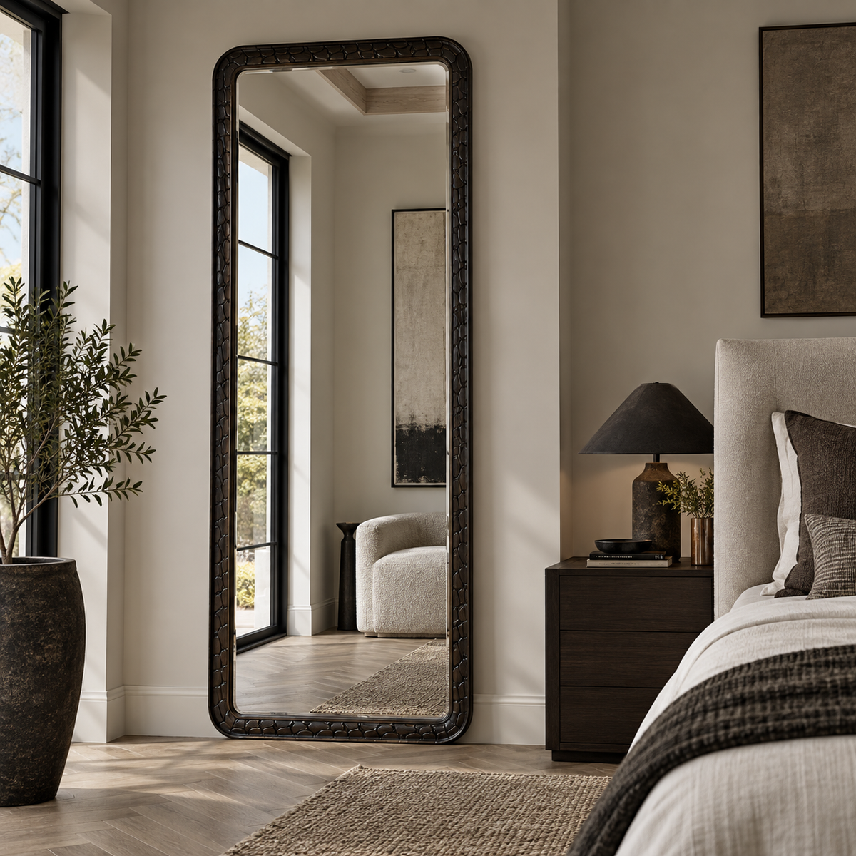 A tall floor mirror with an ornate dark frame stands in a styled bedroom next to a bed and nightstand, reflecting a softly lit interior.
