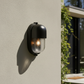 A modern black outdoor wall sconce with a clear ribbed glass shade, mounted on a textured beige stucco wall and casting a warm light, with a blurred outdoor background on the right.