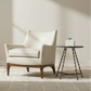 A cream-colored leather armchair with a wooden base sits on a light rug next to a small round side table in a bright, modern room with natural sunlight.