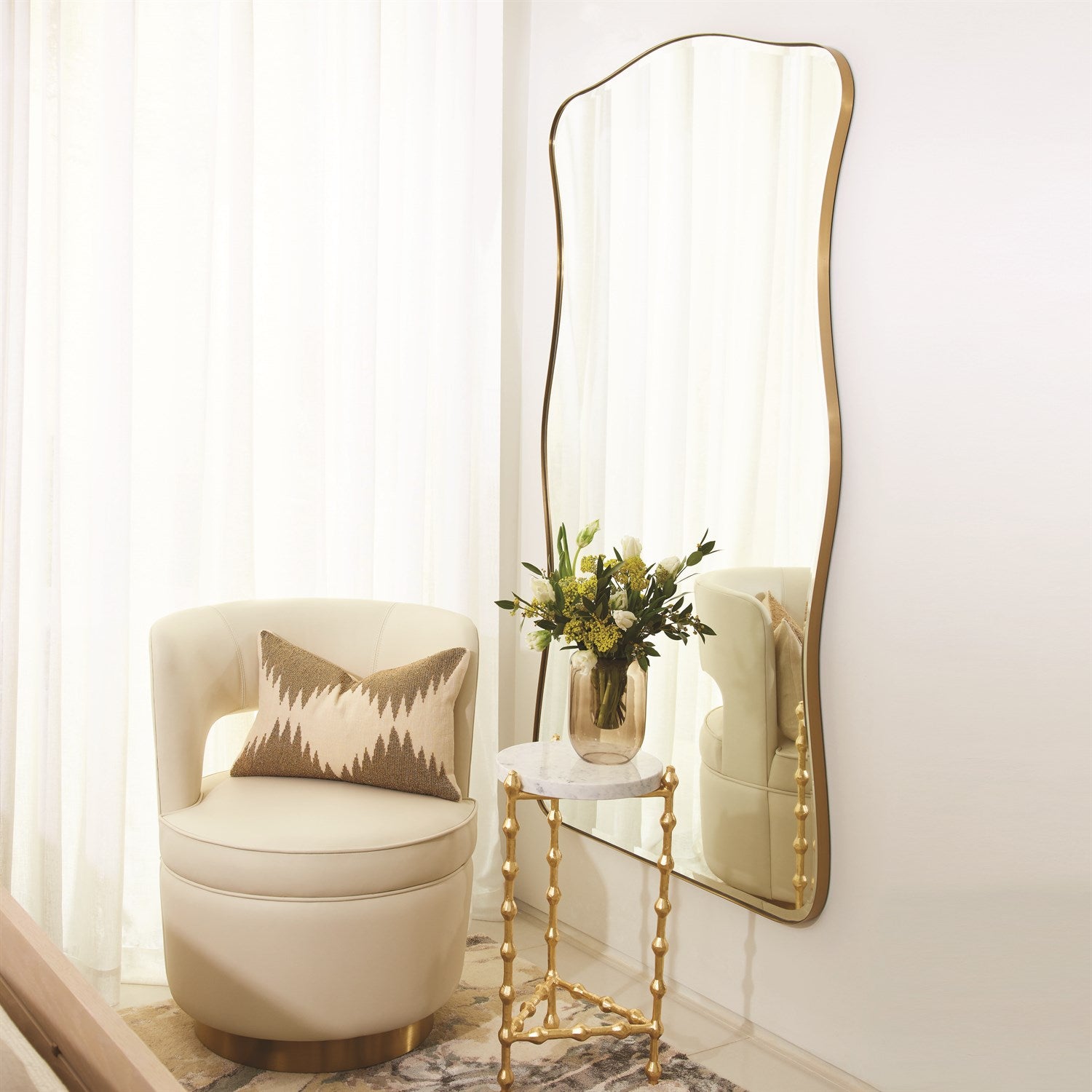 Beige armchair with a decorative pillow next to a tall gold-framed mirror on a white wall.