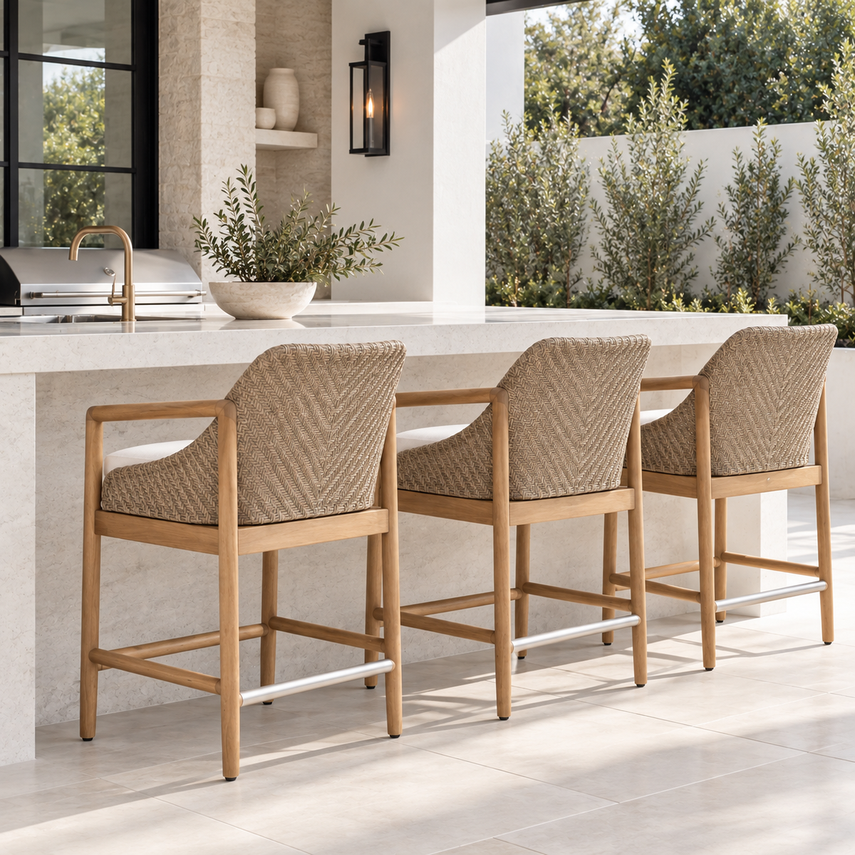 Three natural woven counter stools with white cushions styled at a bright, high-end outdoor patio bar with white stone finishes, black accents, and lush greenery.