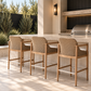Three woven rattan bar stools with natural wood frames and white cushions styled at a modern stone outdoor kitchen bar with black accents and lush landscaping.