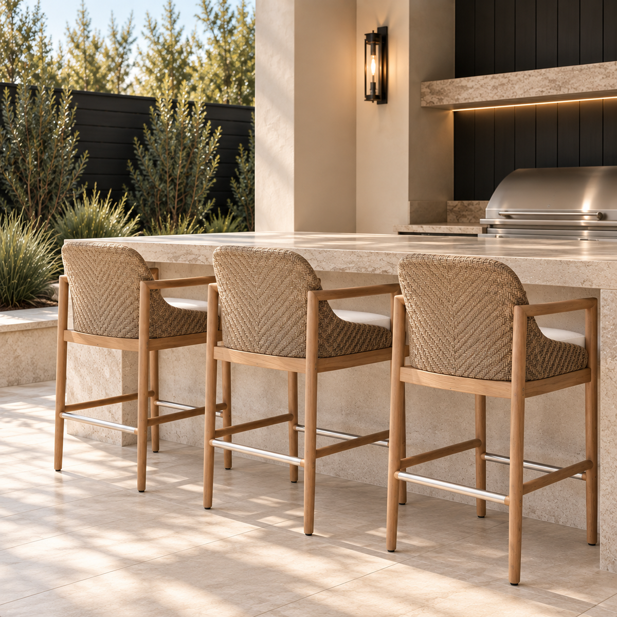 Three woven rattan bar stools with natural wood frames and white cushions styled at a modern stone outdoor kitchen bar with black accents and lush landscaping.