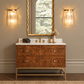 A luxury bathroom vanity featuring a rich burl wood cabinet with brass hardware, a white marble countertop, and a matching gold-framed mirror. Flanking the mirror are two matching gold wall sconces, and the scene is completed with a woven rug on a dark wood floor.