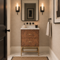 A luxury bathroom vanity featuring a copper-brown textured cabinet with gold legs, a white marble countertop, and a large rectangular mirror above. The bathroom has beige wallpaper, black sconces, a black door on the left, and a white towel hanging on the right.