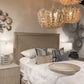 A styled bedroom scene featuring a bed with white linens, blue and beige accent pillows, a textured beige upholstered headboard, matching nightstands, lamps, and two large leaf-shaped pendant chandeliers hanging above.