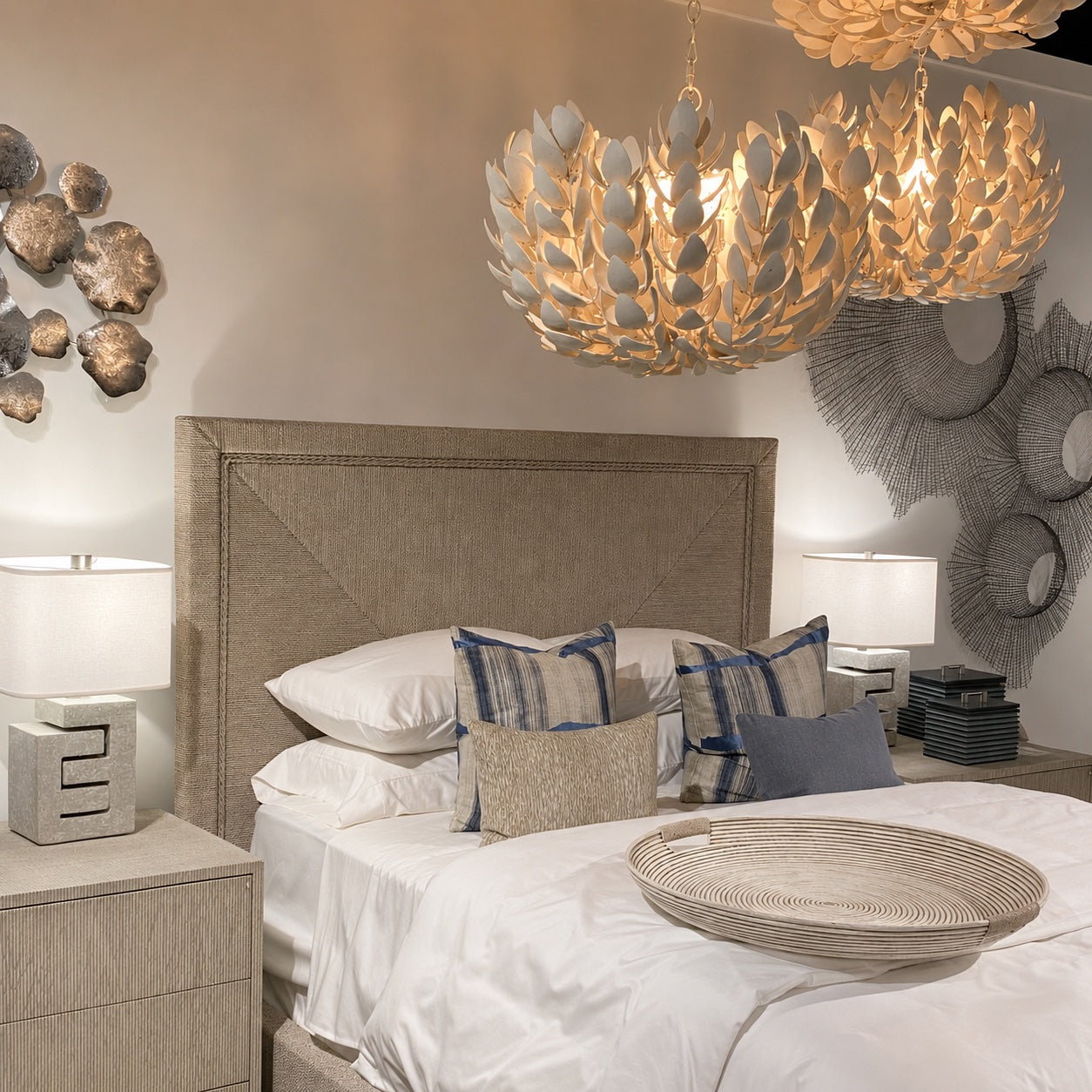 A styled bedroom scene featuring a bed with white linens, blue and beige accent pillows, a textured beige upholstered headboard, matching nightstands, lamps, and two large leaf-shaped pendant chandeliers hanging above.