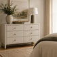 A white six-drawer dresser in a beautifully styled bedroom, featuring small gold knobs, a lamp, a vase with greenery, and a framed landscape print. The bed with beige linens and a dark green throw blanket is visible on the right, with soft window light and curtains in the background.
