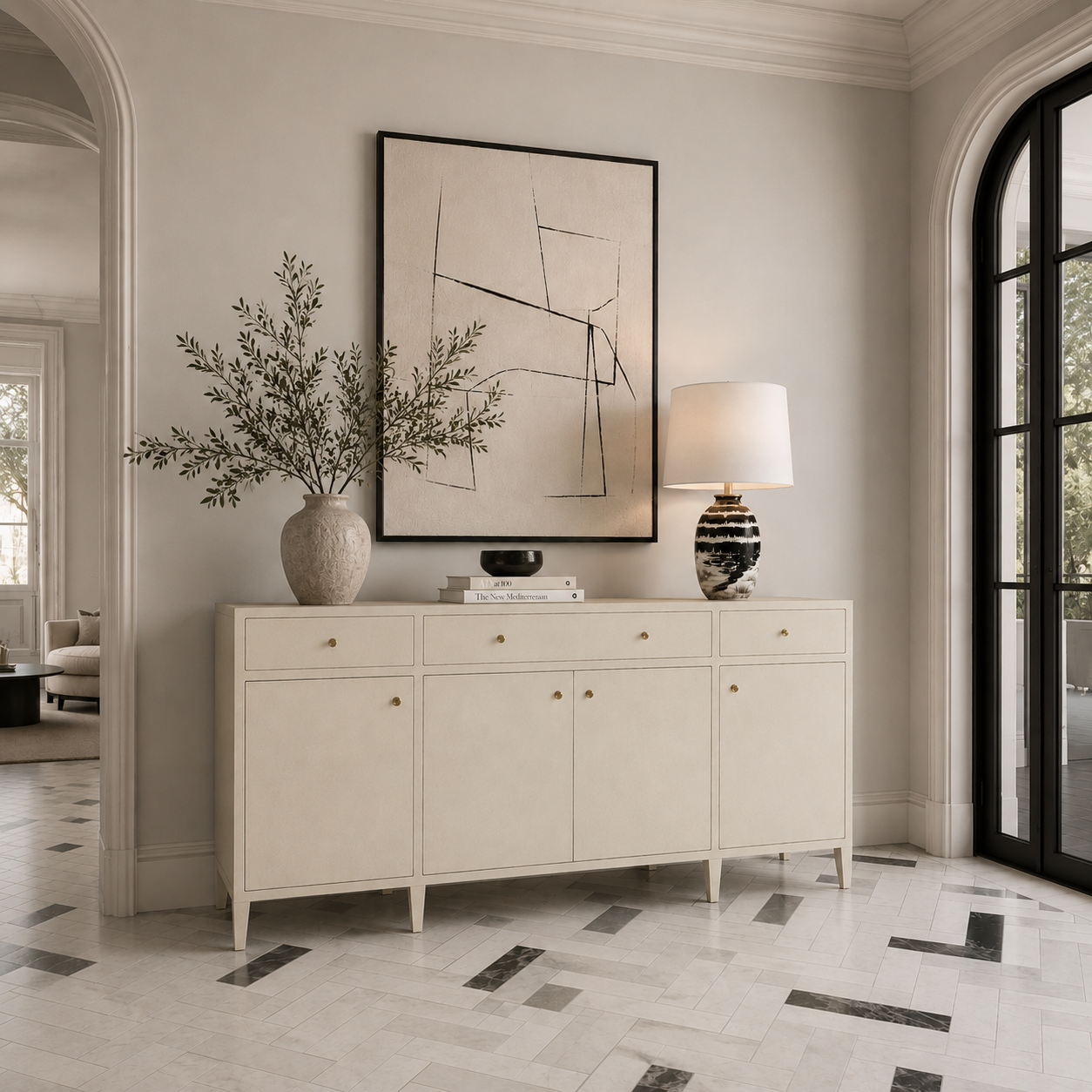 A cream-colored wooden console cabinet with gold knobs, styled in a bright, elegant entryway with a vase, books, a lamp, and a large framed abstract print on a tiled floor.