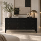 A black wood dresser with six drawers and small brass knobs, styled in a modern living room setting with a vase, books, a ring light, and a lamp on top.