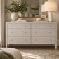 A light beige linen-textured dresser with six drawers and small gold knobs, styled in a bedroom setting with a lamp, books, a vase with greenery, and a mirror in the background.