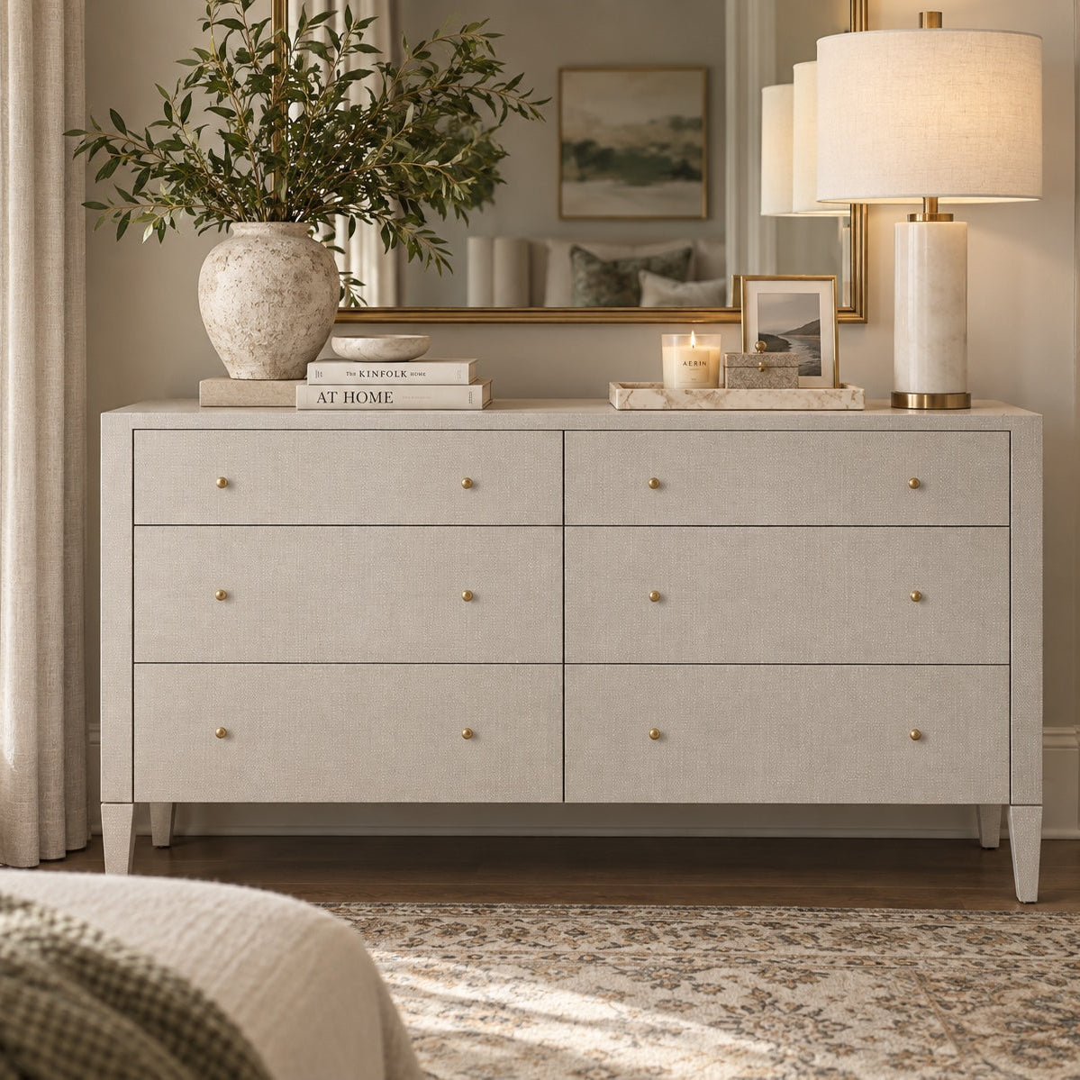 A light beige linen-textured dresser with six drawers and small gold knobs, styled in a bedroom setting with a lamp, books, a vase with greenery, and a mirror in the background.