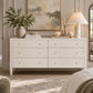 An off white six-drawer dresser with small gold knobs in a beautifully styled bedroom setting, decorated with a vase of greenery, candles, and a lamp.