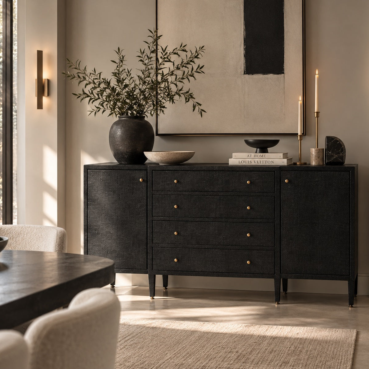 A black wood sideboard or credenza with brass drawer knobs, styled in a modern dining room setting with a vase of greenery, candles, and wall sconces.