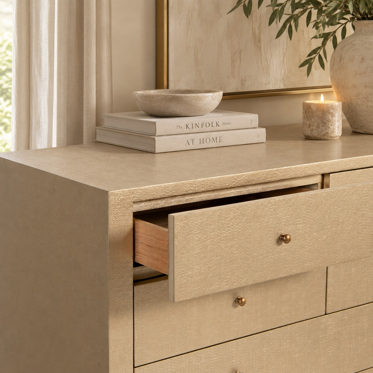 A gold/beige textured dresser with an open drawer, styled with books, a bowl, a candle, and a vase in a warm, elegant room setting.