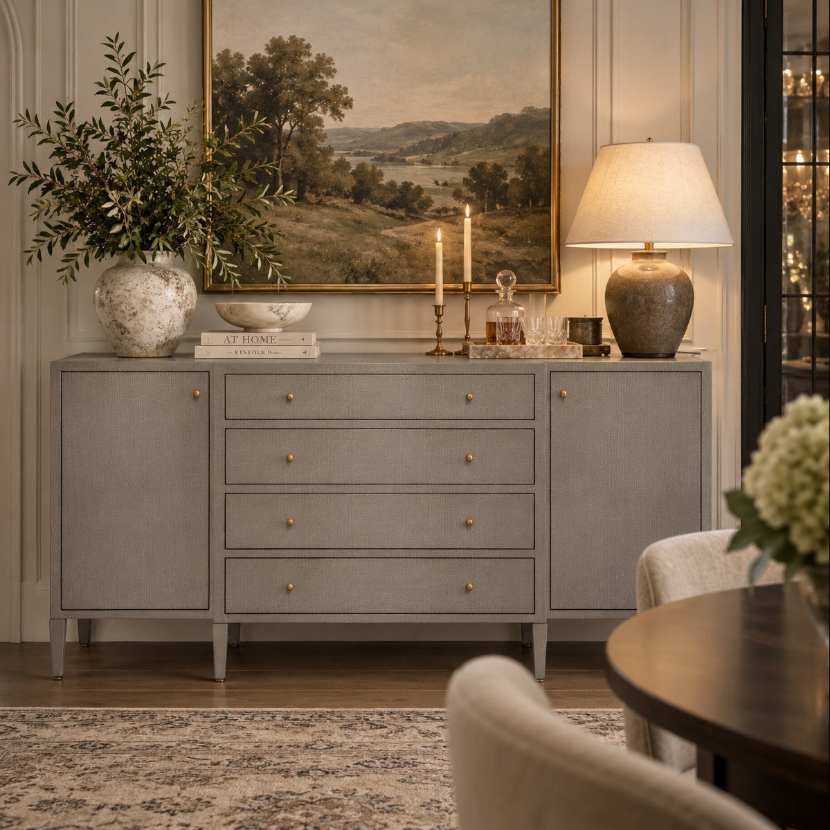 A gray sideboard console cabinet with five drawers and brass knobs, styled in a classic elegant room with a lamp, vase, and a large landscape painting on the wall.