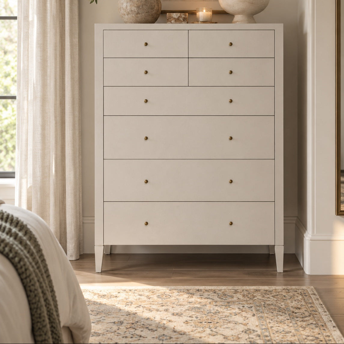 A tall light gray wooden chest of drawers with six drawers and small round brass knobs, styled in a bright bedroom setting with a vase, candle, and bowl on top.