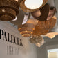 A collection of woven and wicker pendant light fixtures in various organic, sculptural shapes hanging from a ceiling in a showroom. The lights feature natural brown and beige woven materials, with one prominent white flower-like chandelier in the foreground.