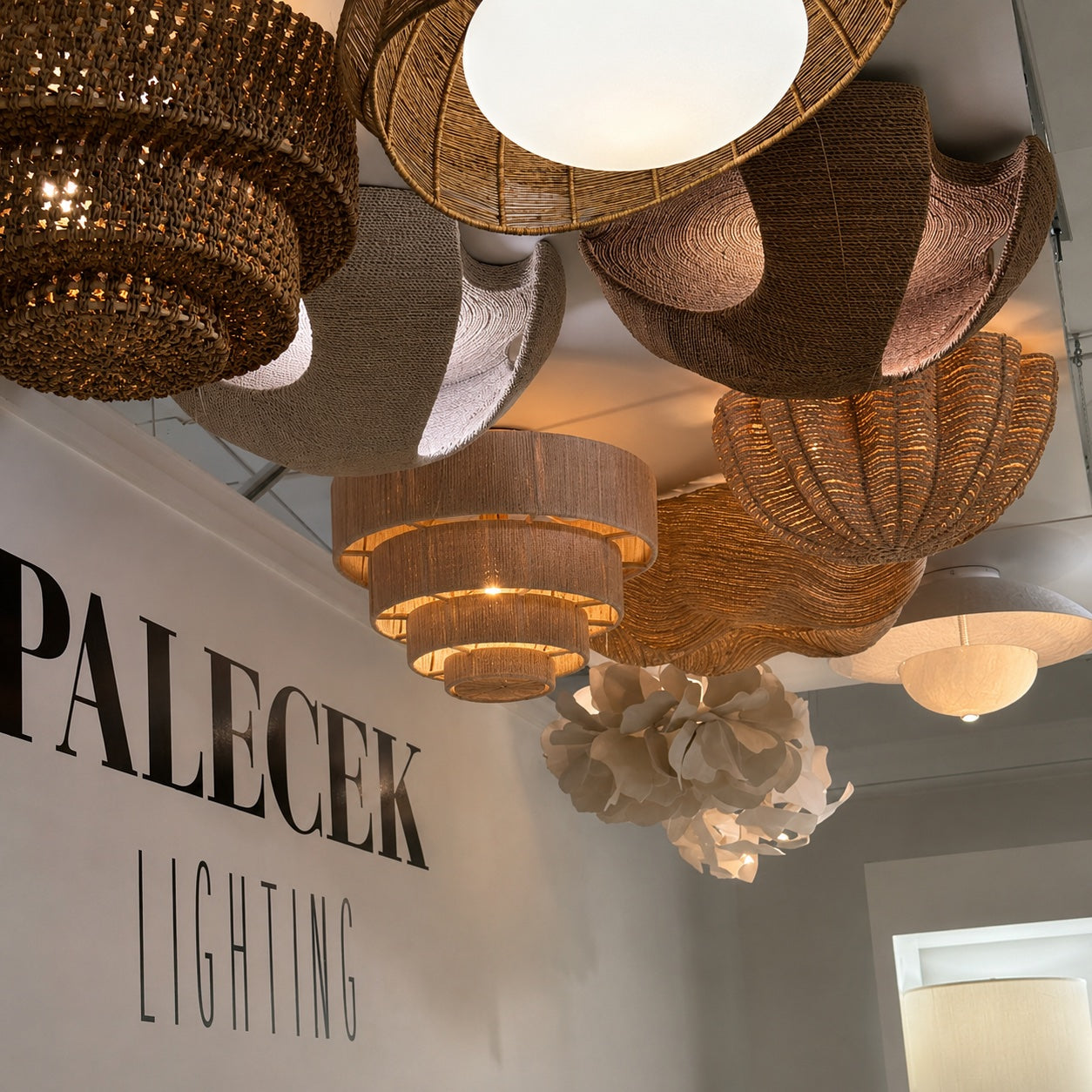 A collection of woven and wicker pendant light fixtures in various organic, sculptural shapes hanging from a ceiling in a showroom. The lights feature natural brown and beige woven materials, with one prominent white flower-like chandelier in the foreground.