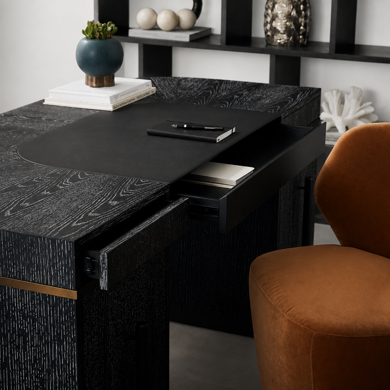 A modern black desk with a textured wood grain finish, featuring an open drawer and a matching black desk mat. The desk is styled in a home office setting with a brown velvet chair, books, and decorative objects on a shelf in the background.
