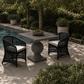 Outdoor patio dining set with a square gray stone-look table and two black woven armchairs with white cushions, placed beside a swimming pool in a landscaped garden.