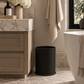 A black woven cylindrical wastebasket bin placed on a marble-tiled bathroom floor next to a vanity and bathtub.