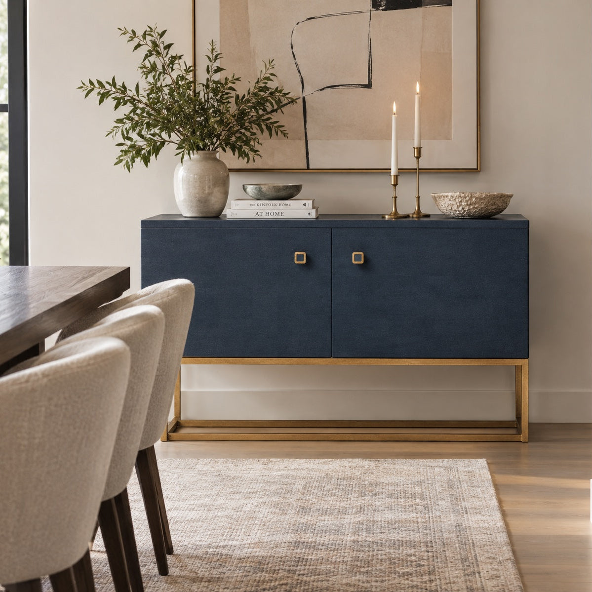 A navy blue sideboard cabinet with a gold metal frame and square gold knobs, styled in a dining room setting with a vase of greenery, candles, and books on top.
