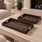 Two dark brown textured rectangular serving trays with gold linear accents and side handles, displayed on a light wood coffee table in a modern living room setting.