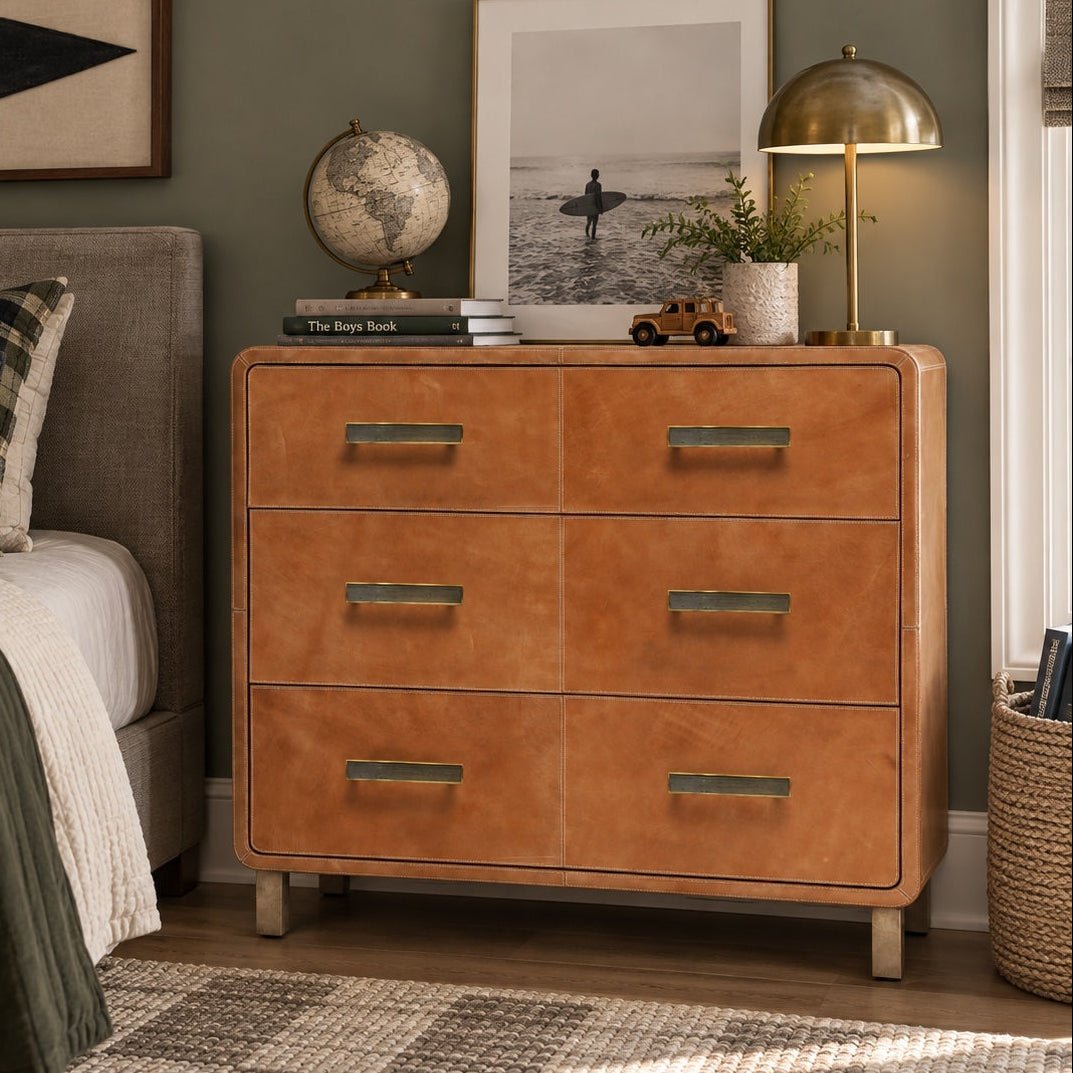 A three-drawer wooden dresser with warm tan leather-look fronts and brass handles, styled in a bedroom next to a bed with a gray headboard, framed wall art, a globe, and a brass table lamp.