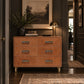 A warm tan leather six-drawer dresser with metallic gold handles, styled in a dark hallway with a lamp, framed landscape print, and rug.