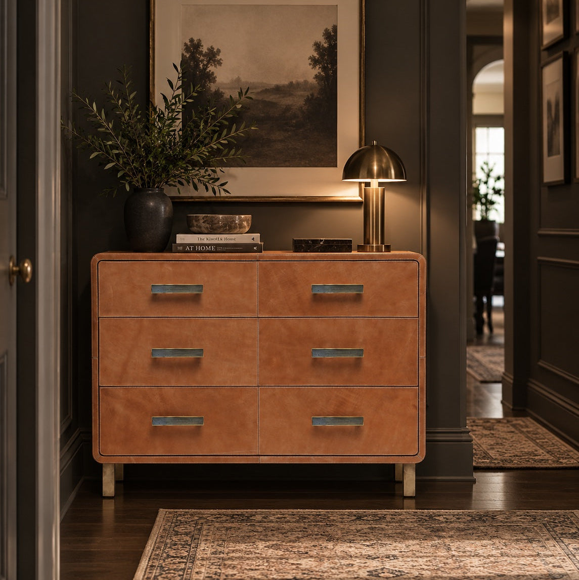A warm tan leather six-drawer dresser with metallic gold handles, styled in a dark hallway with a lamp, framed landscape print, and rug.