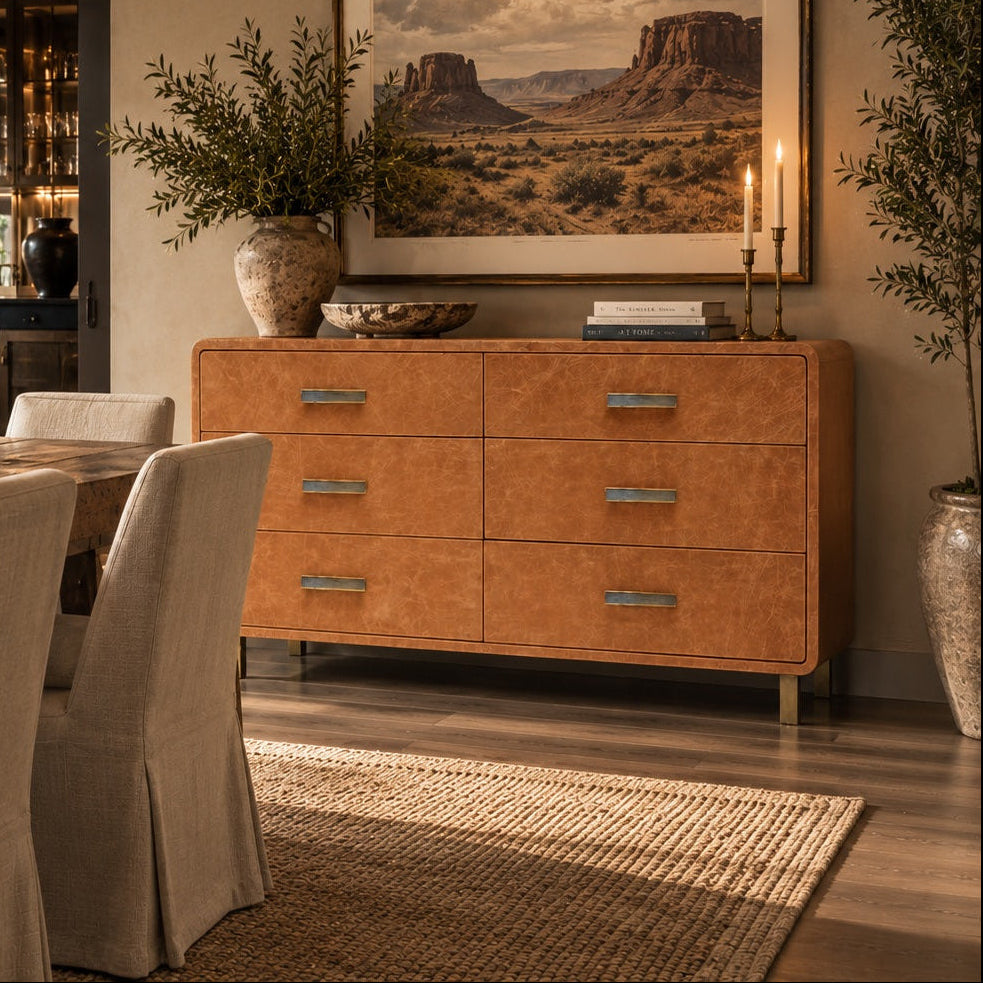 A styled dining room interior featuring a warm orange-brown wooden sideboard with silver handles, decorated with a large framed landscape painting of Monument Valley, candles, and plants.