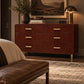 A dark reddish-brown leather six-drawer dresser in a styled bedroom setting, topped with books, a vase of greenery, and a lit brass table lamp under a framed landscape painting.