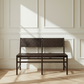 A dark brown bench with woven rope seats and backs, positioned against a white paneled wall with soft sunlight casting shadows.