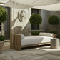 A modern outdoor daybed with a light wood frame and white cushions, styled on a stone patio under a white canopy with potted plants.