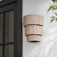 A three-tiered cylindrical outdoor wall sconce with a natural beige woven rope texture, mounted on a white brick wall next to a black-framed glass door.