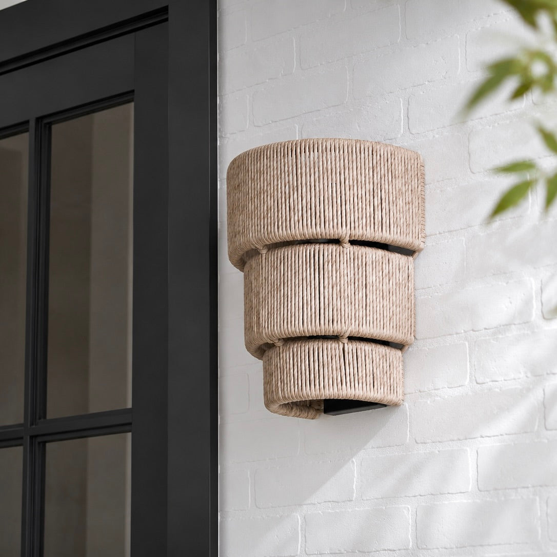 A three-tiered cylindrical outdoor wall sconce with a natural beige woven rope texture, mounted on a white brick wall next to a black-framed glass door.