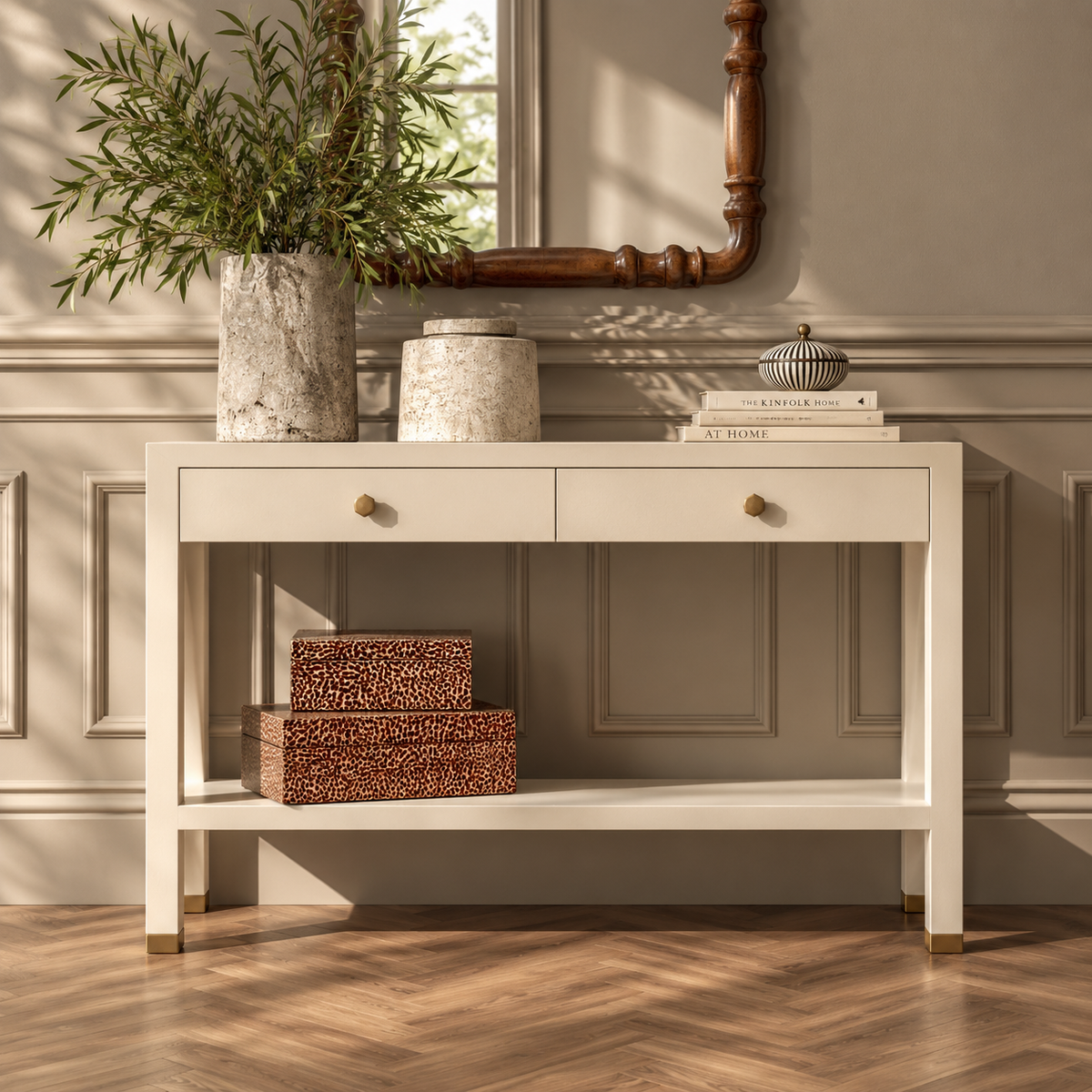 A white console table with two drawers and an open lower shelf, styled in a warm, sunlit room with wood paneling, a mirror, books, and decorative boxes.