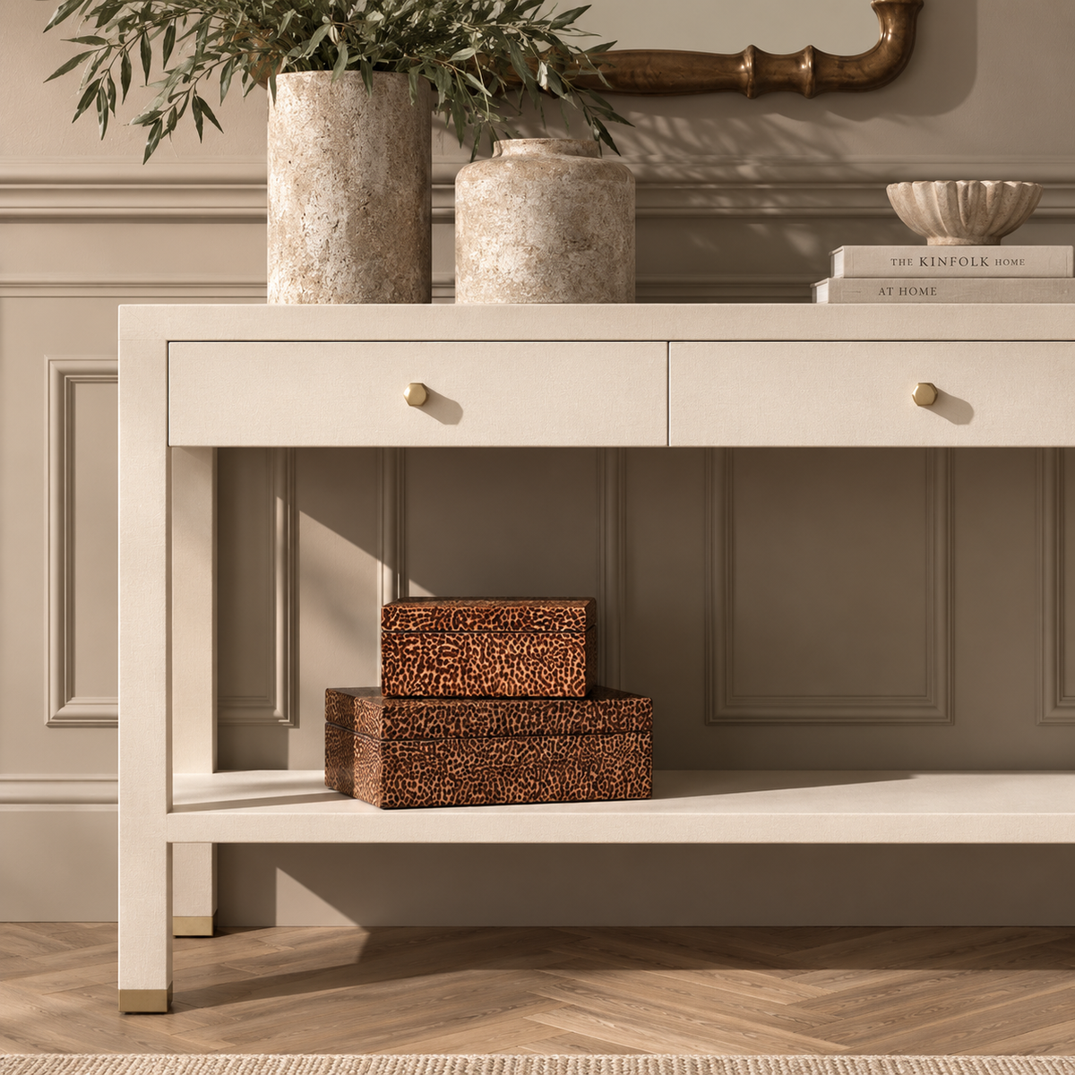 A cream-colored console table with two drawers and an open lower shelf, styled in a home entryway with decorative vases, books, and storage boxes. The table features small brass knobs and gold-toned metal feet, set against a neutral paneled wall and wood flooring.