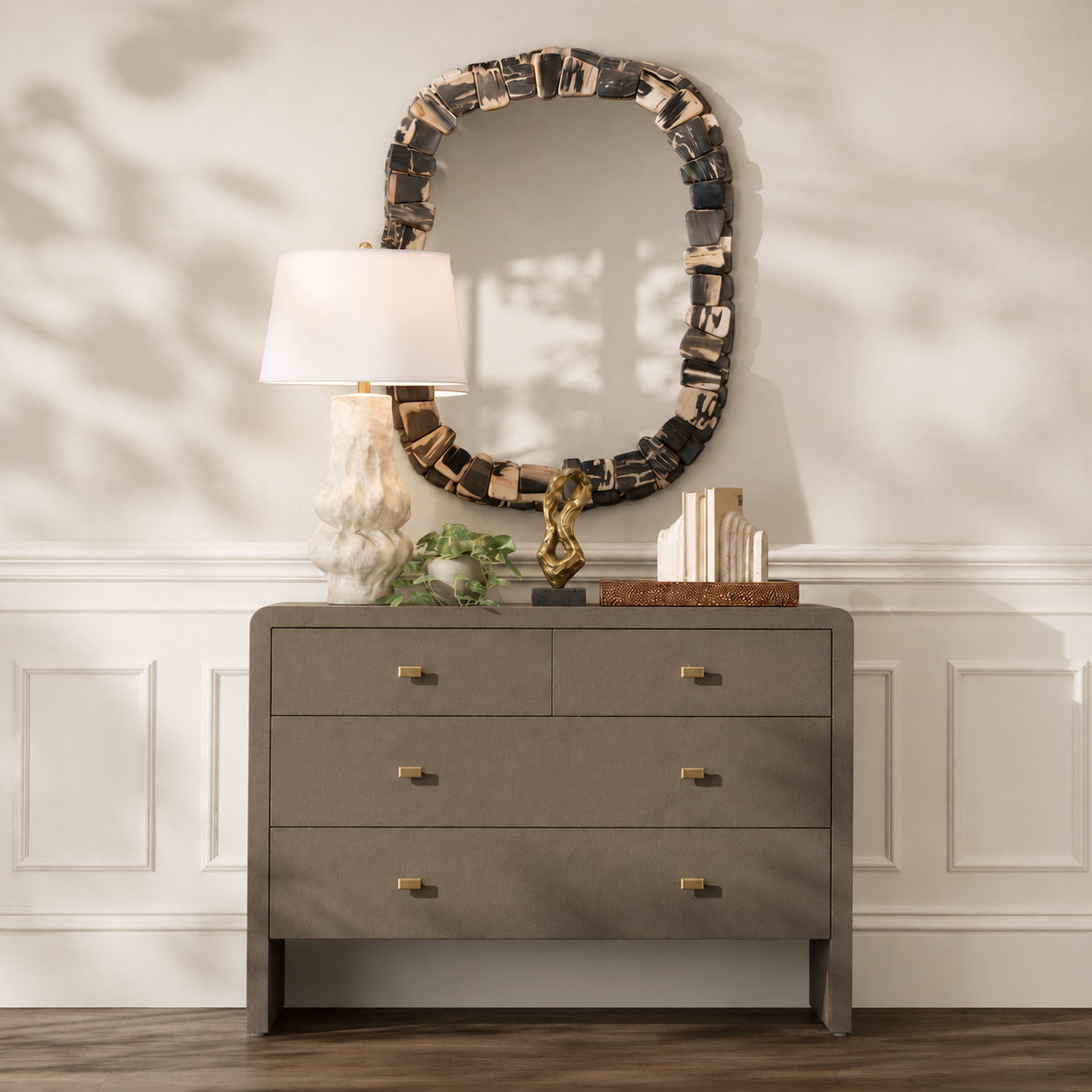 A gray three-drawer dresser with brass handles in a styled room setting, topped with a white lamp, a decorative oval mirror, and small plants.