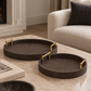 Two round dark brown woven tray coasters with gold metal corner accents resting on a light marble coffee table in a modern living room setting.