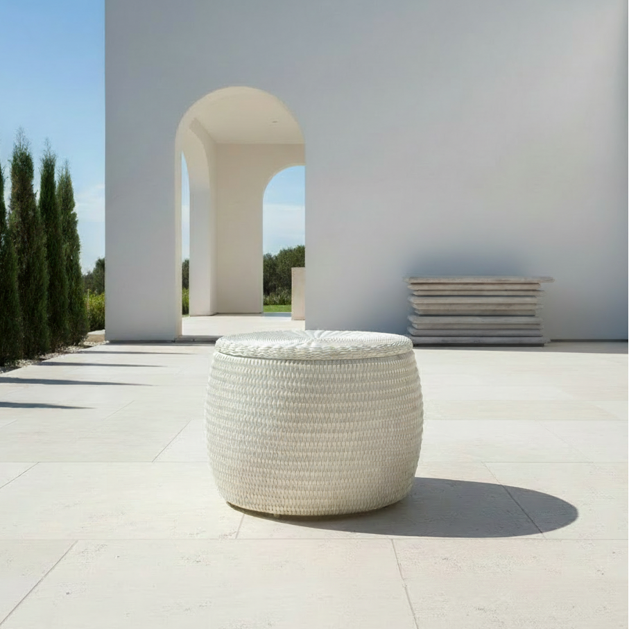 A round, white woven outdoor pouf or ottoman with a matching round lid, set on a light stone patio in front of a modern white architectural structure with an arched opening.